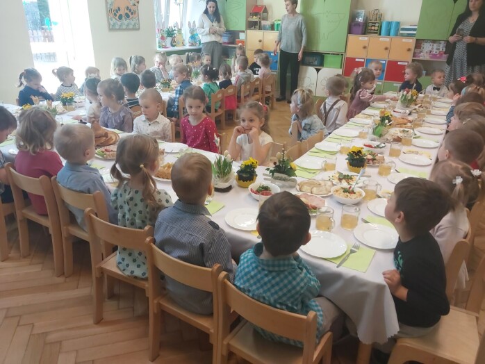 dzieci odświętne ubrane siedzą przy nakrytym duzym stole wielkanocnymi smakołykami