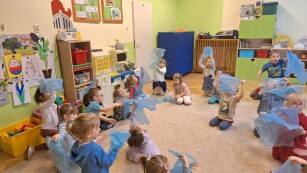 dzieci siedzą w kółku na dywanie w kolorowej sali trzymając nad głową niebieskie worki