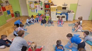 dzieci siedzą w kółku na dywanie w kolorowej sali tzrymając przed sobą worki pani tłumaczy zabawę