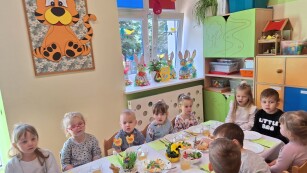 widok części stołu grupka dzieci siedzi przy odświętnie nakrytym stole z białym obrusem i dekoracjami wielkanocnymi zza okien przebija słońce i oświetla pomieszczenie na parapecie stoją kolorowe zające zrobione z kartonu