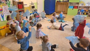 dzieci siedzą w kółku na dywanie w kolorowej sali jedno dziecko w środku kółka leży na brzuchu
