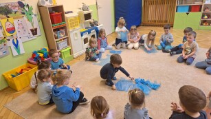 dzieci siedzą w kółku na dywanie w kolorowej sali jeden chłopiec na środku kółka sięga po niebieski worek leżący na dywanie