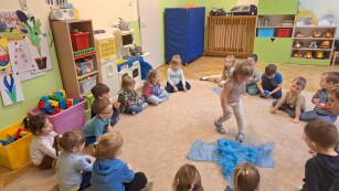 dzieci siedzą w kółku na dywanie w kolorowej sali jedna dziewczynka na środku kółka sięga po niebieski worek leżący na dywanie