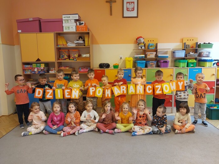 grupa dzieci ustawiona do zdjęcia z napisem Dzień Pomarańcowy, dzieci stojące trzymają napis, pozostałe dzieci siedzą na dywanie