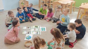 Dzieci siedzą na dywanie w kręgu a w środku znajdują się ilustracje do zajęć z okazji Dnia Ziemi