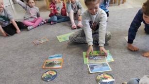 Dzieci siedzą na dywanie na którym rozłożone są obrazki