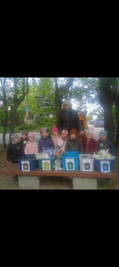 grupa dzieci pozuje do zdjęcia, wśród dzieci jest pani, na tle dzieci - na ławce ustawione sa pojemniki z etykietami dotyczącymi segregacji odpadów