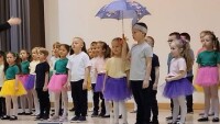 grupa dzieci kolorowo ubranych występuje na scenie śpiewając, jeden chłopiec trzyma w ręku rozłożoną parasolkę w kolorze fioletowym