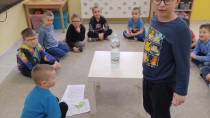 dzieci siedzą na dywanie dookoła białego stolika na którym stoi butelka z wodą jeden chłopiec stoi patrząc w obiektyw