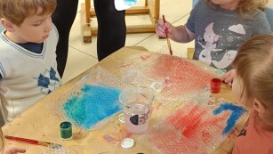 Dzieci maluja farbami, pośród nich pani prawdopodobnie objaśniająca zadanie do wykonania
