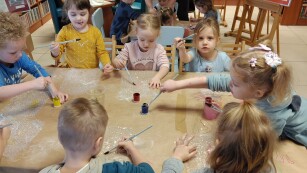 Dzieci siedzą przy stole, wokół książki. Dzieci malują farbami.
