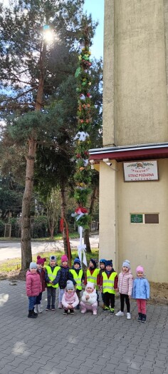 grupa dzieci stoi przy wysokiej palmie, w tle budynek OSP