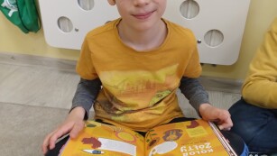 chłopiec w okularach ubrany w pomarańczową bluzę trzyma na kolanach otwartą książkę