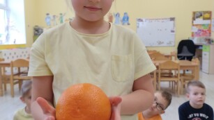 dziewczynka o blond włosach ubrana w żółtą koszulkę trzyma w rękach pomarańczowy owoc uśmiecha się