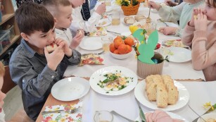zbliżenie na długi stół przy którym jedzą dzieci na stole biały obrus i nakrycia z potrawami świątecznymi panie pomagają nakładać jedzenie
