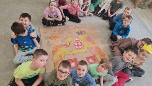 zdjęcie z góry dzieci siedzą w kółku patrząc w obiektyw po środku leży szary dyży karton z rysunkiem dwóch królów i herbu Polski