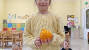 dziewczynka o ciemnych włosach ubrana w żółtą bluzę trzyma w rękach pomarańczowy owoc