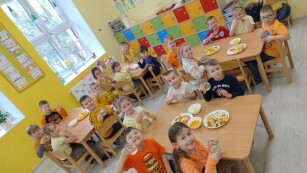 dzieci ubrane w żółte i pomarańczowe ubrania siedzą przy stolikach w przedszkolu w miseczkach stoją pomarańczowe owoce