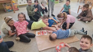 dzieci siedzą na dywanie na środku duża szara kartka czworo dzieci leży na kartce dzieci zerkają w obiektyw trzymają kartki z godłem Polski