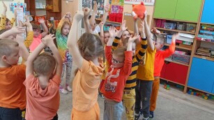 dzieci stoją na dywanie z uniesionymi rękami, bawią się w podawanie piłki