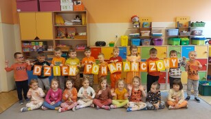 grupa dzieci ustawiona do zdjęcia z napisem Dzień Pomarańcowy, dzieci stojące trzymają napis, pozostałe dzieci siedzą na dywanie