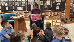 Dzieci siedzą na dywanie w bibliotece. Pani bibliotekarka prezentuje im ilustrację.