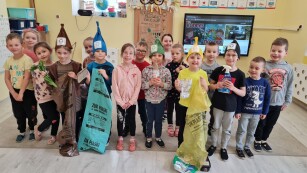 grupa dzieci w sali przedszkolnej stoi trzymając worki do segregacji śmieci,  na głowie dzieci mają wysokie czapki w kolorach brązu i zieleni